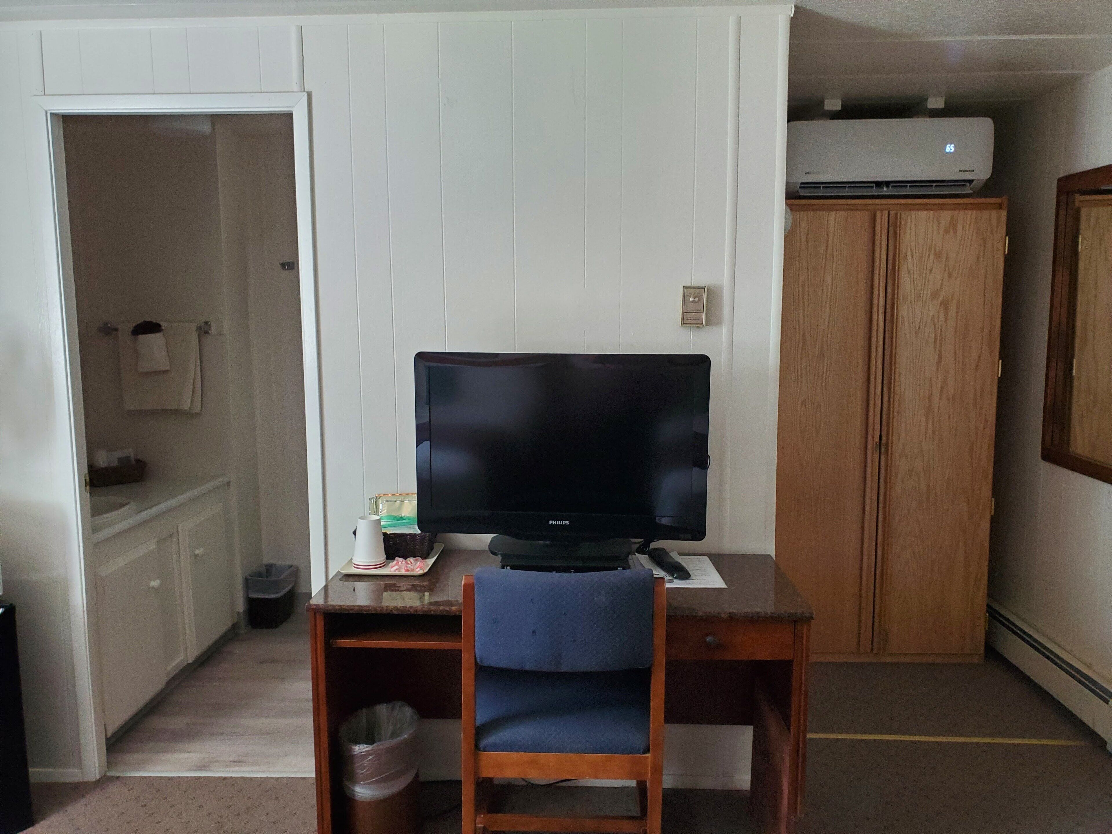 Een kamer met een houten bureau, een televisie, een prullenbak en een houten garderobe.