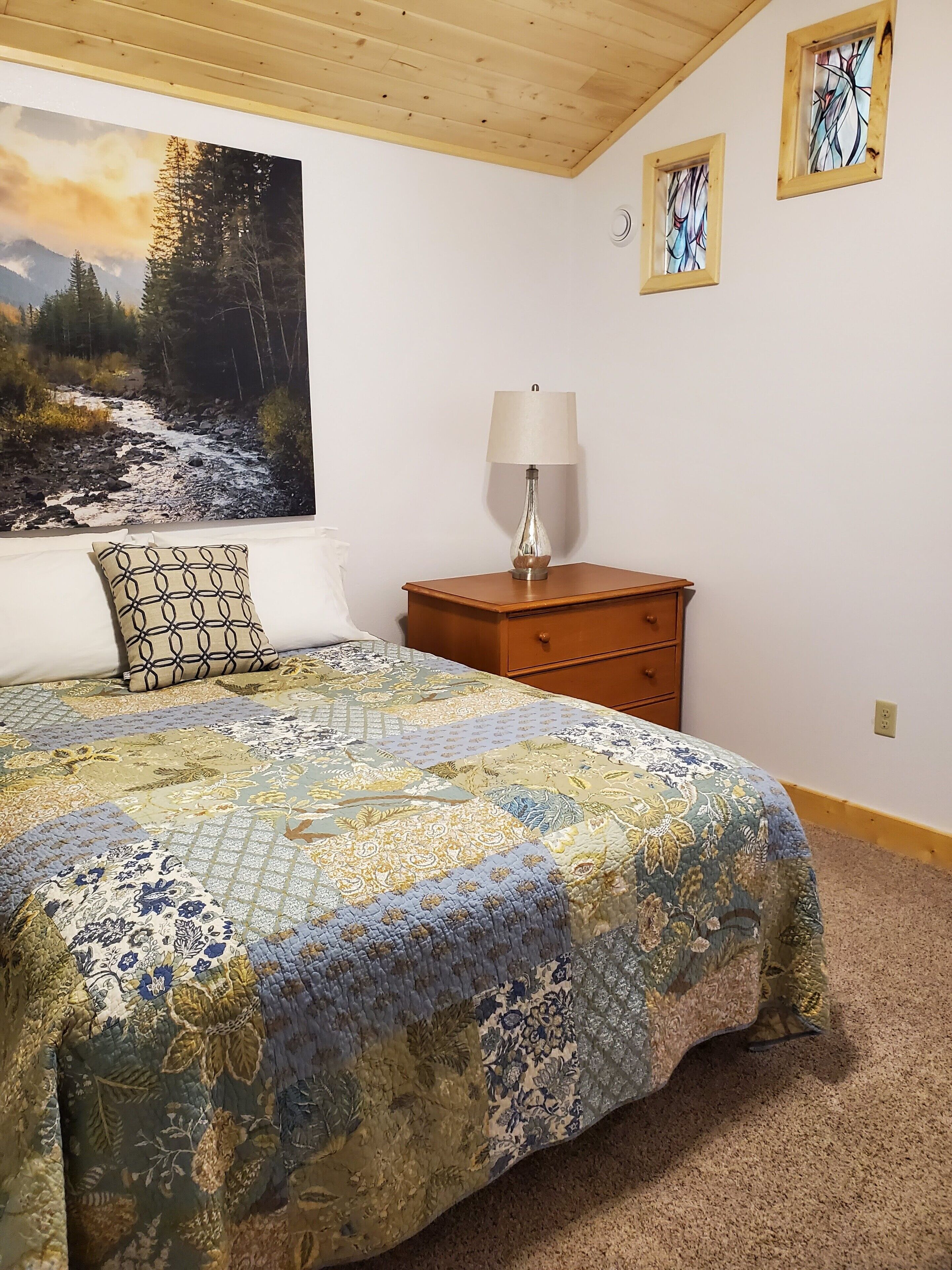 A bedroom with a large bed, a nightstand with a lamp, a dresser, and a painting of a forest stream on the wall.