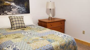 A bedroom with a large bed, a nightstand with a lamp, a dresser, and a painting of a forest stream on the wall.