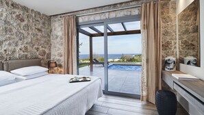 Ein Schlafzimmer mit einem großen Bett, einem Nachttisch, einer Lampe, Blick auf das Meer und einem Badezimmer mit Waschbecken und Spiegel.