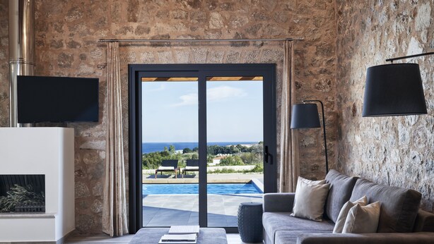 Ein modernes Wohnzimmer mit Steinwand, Kamin, einer Couch und Blick auf Pool und Landschaft.