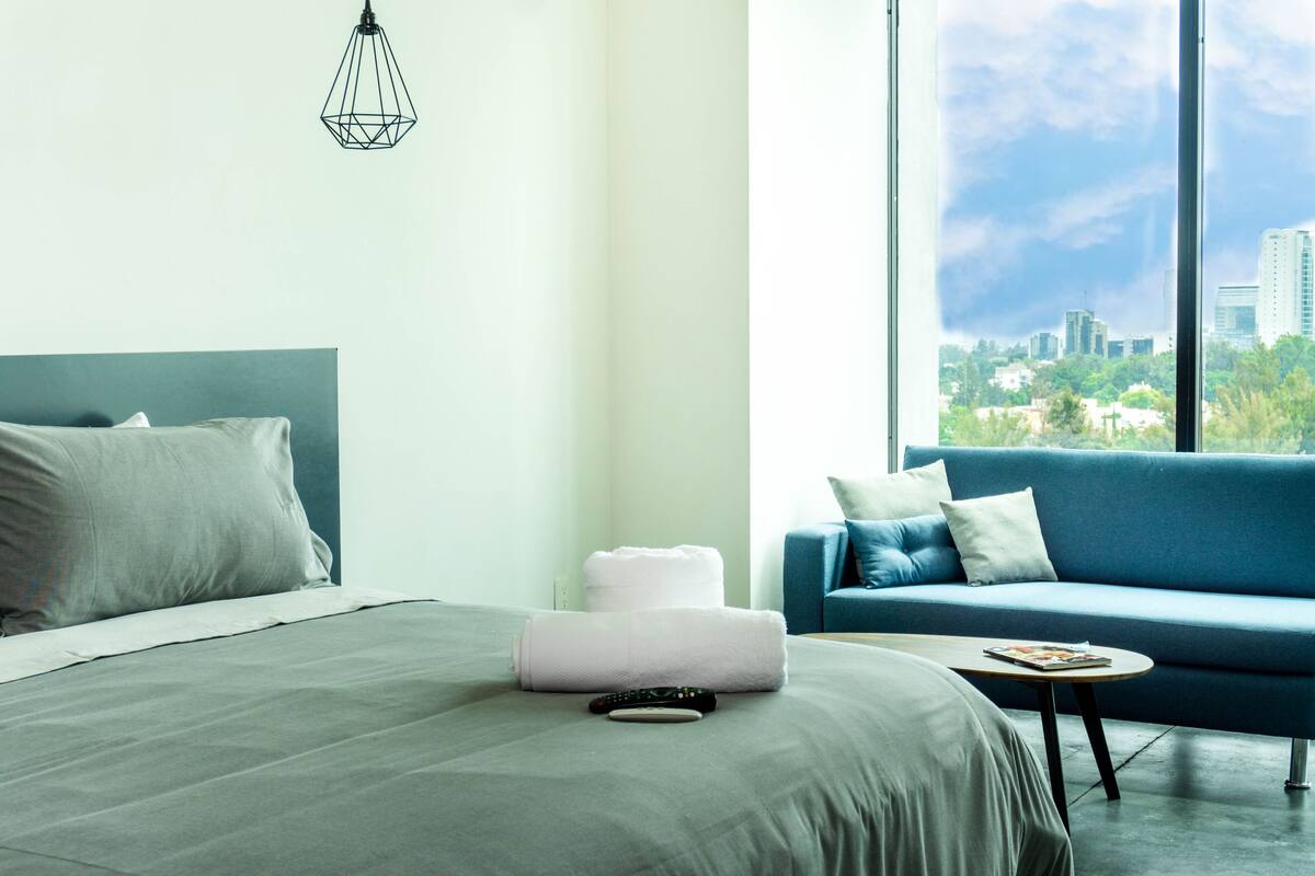 Ein Hotelzimmer mit Bett, blauem Sofa, kleinem Tisch und Blick auf eine Stadtkulisse durch große Fenster.