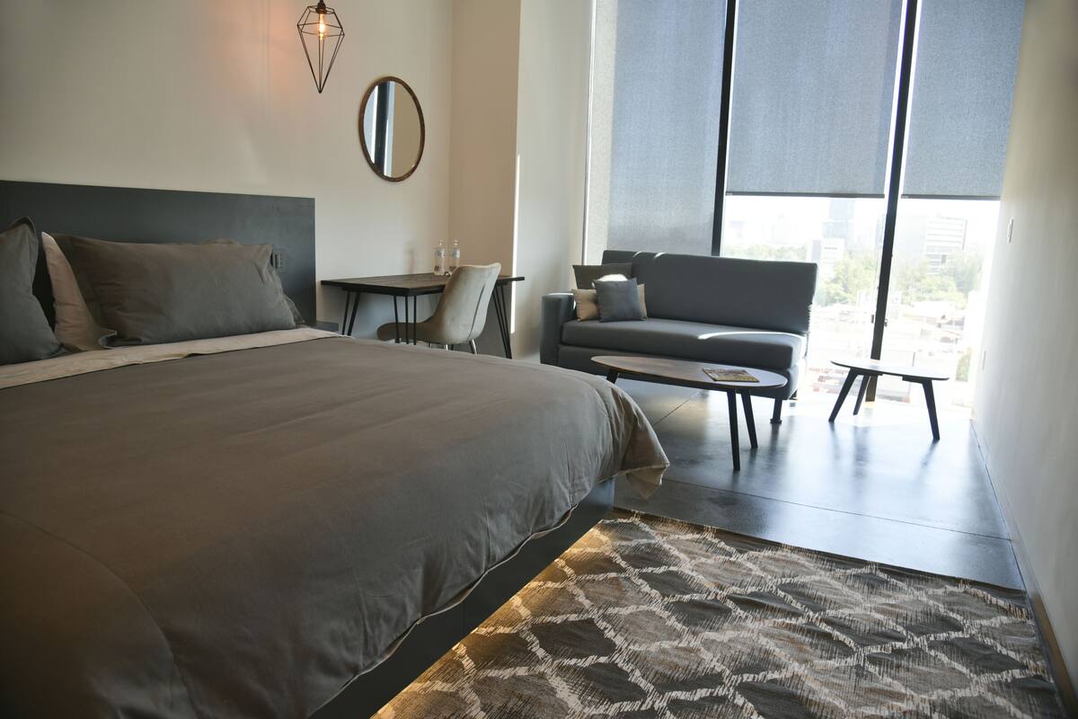Ein modernes Hotelzimmer mit einem großen Bett, einer Couch, einem kleinen Tisch und einem Sessel. Ein Fenster mit Jalousien, ein Spiegel an der Wand und ein gemustertes Teppich sind ebenfalls vorhanden.