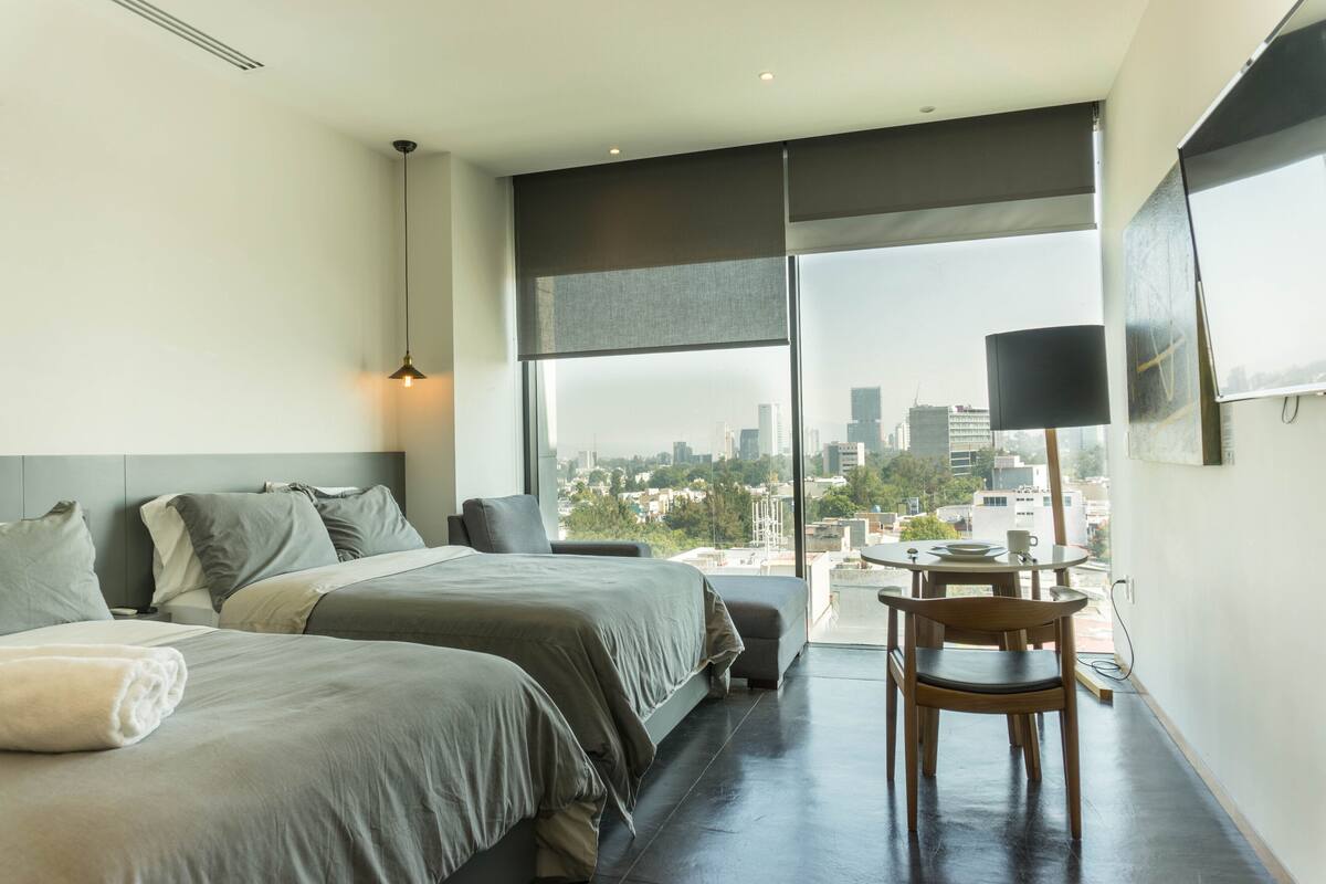 Ein modernes Hotelzimmer mit zwei Betten, einem Esstisch und einem großen Fenster mit Blick auf die Stadt.