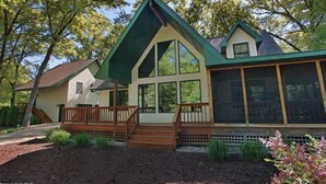 A two-story house with a green roof, a front porch with a wooden railing, and a large window overlooking a garden.