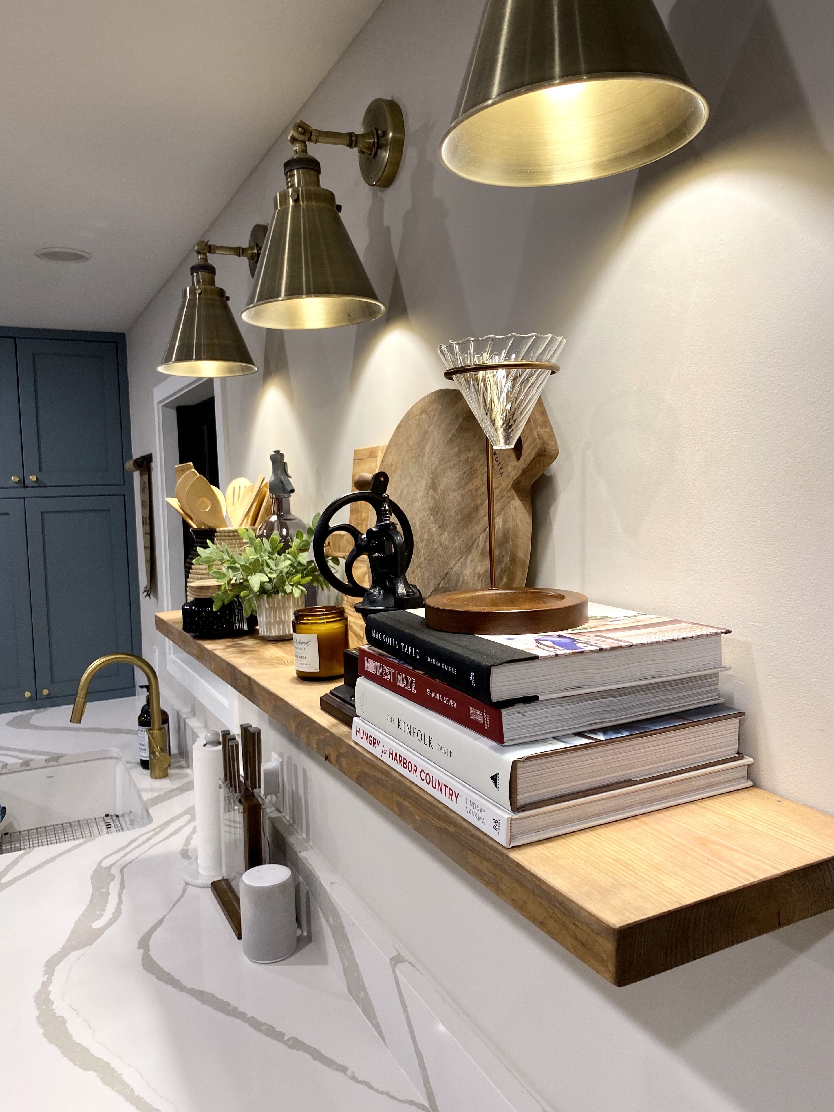 Une cuisine avec un plan de travail en bois, un évier et une machine à café. Des livres sont empilés sur le plan de travail et une plante est accrochée au mur.