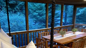 A wooden deck with a table and chairs, a cushioned bench, and large windows offering a view of trees.