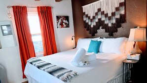 A neatly made bed with white linens, a patterned throw blanket, and two towels. A wall-mounted lamp and a framed picture are visible. - Nob Hill’s Casa Flamingo: King & Queen beds, Safe, Prime Location, Large Yard. (Albuquerque)