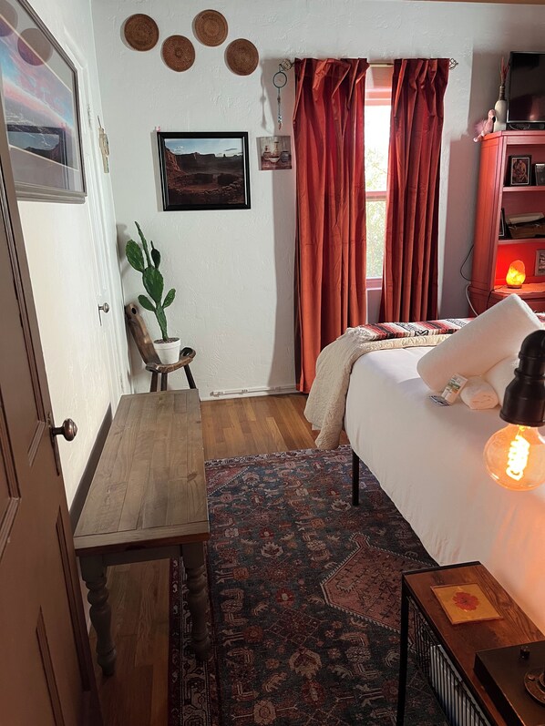 Un dormitorio acogedor con cama, mesita de noche, mesa, silla, un cactus, alfombra y ventana con cortinas.
