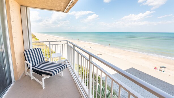 Ein Balkon mit einem gestreiften Stuhl und einem kleinen Tisch, mit Blick auf einen Strand und klares Wasser. In der Ferne sind einige Personen zu sehen.