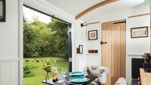 Un comedor con una mesa lista para dos personas, una silla y vistas a un jardín verde con animales.