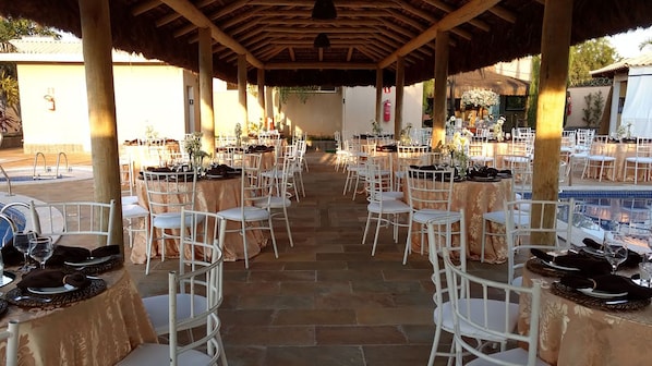 An outdoor covered dining area with multiple round tables, white chairs, and a pool in the background.
