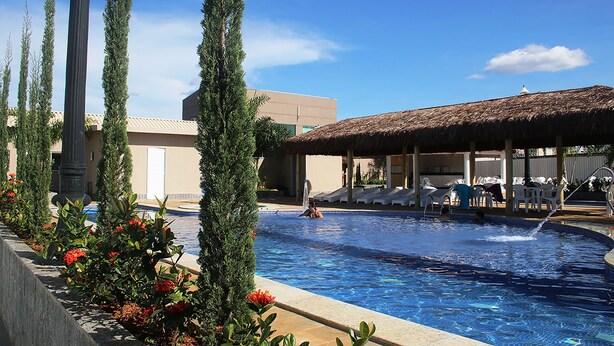 Ein Pool mit überdachtem Bereich, einem Reetdach, umgeben von hoch aufragenden Zypressen und einem Gebäude mit sandfarbenem Mauerwerk.