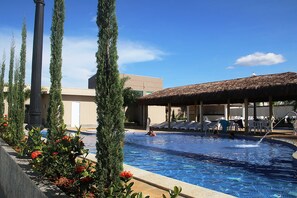 A swimming pool with a thatched roof area, surrounded by tall cypress trees and a building with a beige facade.