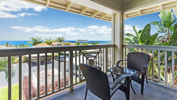 A balcony with wicker chairs and a glass table, overlooking a parking area and the ocean.