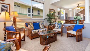 A cozy living area with wicker furniture, a glass coffee table, and a view of a house through large windows.
