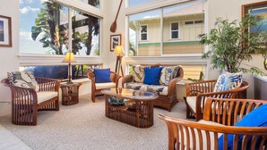 A sunlit room with wicker furniture, a glass coffee table, and a view of a house through large windows.