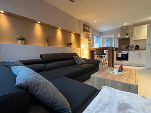 Un salon moderne avec un canapé foncé, une table basse en bois et une cuisine en arrière-plan.