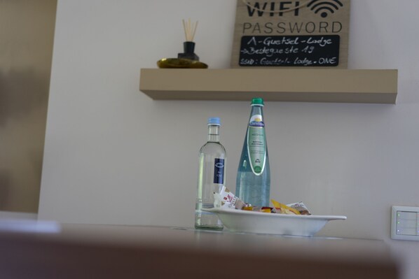 Un comptoir de cuisine avec une assiette de nourriture, une bouteille d’eau et un flacon de gel hydroalcoolique. Une pancarte au-dessus du comptoir indique « Mot de passe WiFi » et « A-Guestel - Lodge Bestgewese st 13 ».