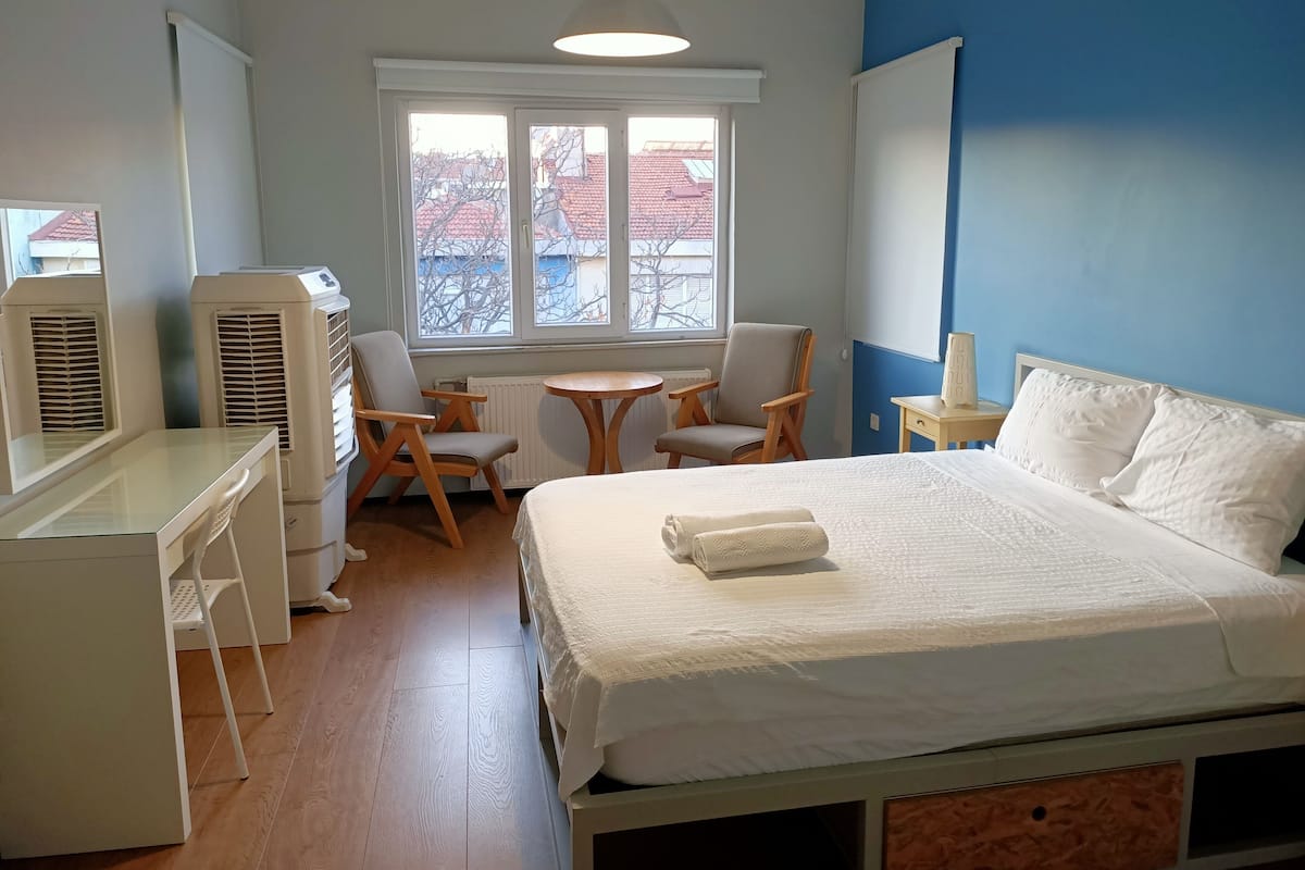 A bedroom with a bed, a desk, two chairs, a small table, and a window with a view of buildings.