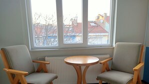 Una habitación con dos sillas de madera, una mesita redonda pequeña, un radiador y una ventana con vistas a edificios.