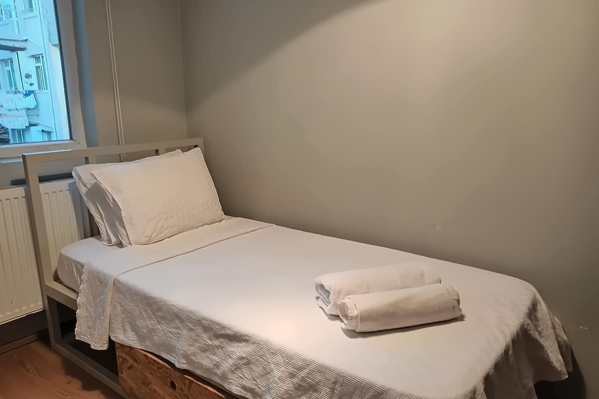 A single bed with a white bedspread and pillows, a wooden headboard, and a window with a view of buildings.