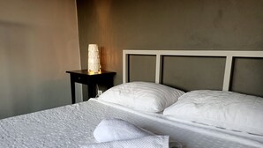 Una cama bien hecha con sábanas y almohadas blancas, una toalla doblada y una lámpara de noche sobre una mesita.