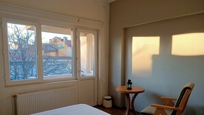 Habitación de hotel con cama, una silla, una mesita con un farolillo y una ventana con vistas a edificios y árboles.