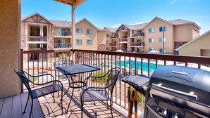 A balcony with outdoor furniture and a grill, overlooking a swimming pool and multi-story buildings. - 5G Perfect Moab RedCliff Condo, POOL AND HOT TUB (Moab)