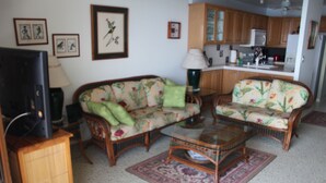 Un salon comprenant un canapé, deux fauteuils, une table basse, un téléviseur et, au fond, un coin cuisine.