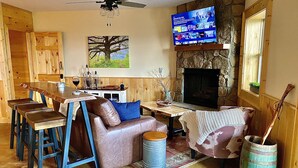 Un salon chaleureux avec une cheminée en pierre, un téléviseur fixé au-dessus, et une table à manger en bois, accompagnée de tabourets de bar.