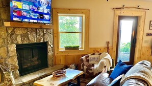 L’intérieur d’un chalet accueillant, avec une cheminée en pierre, un téléviseur fixé au-dessus, un confortable canapé en cuir et une fenêtre donnant sur un paysage verdoyant.