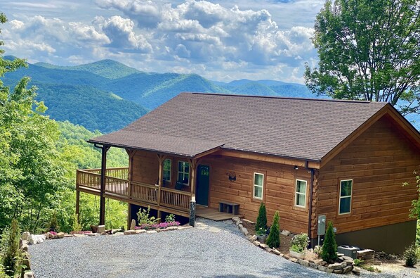 A wooden cabin with a porch, surrounded by a gravel driveway and stone walls, set against a mountainous backdrop.
