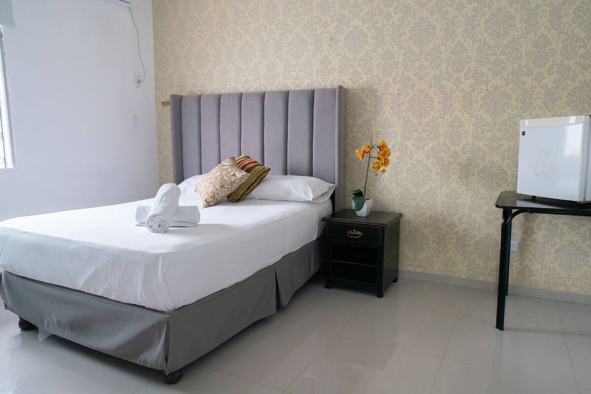A neatly arranged hotel room with a bed, a nightstand with a vase of flowers, a small table with a refrigerator, and a window with a decorative iron grille.