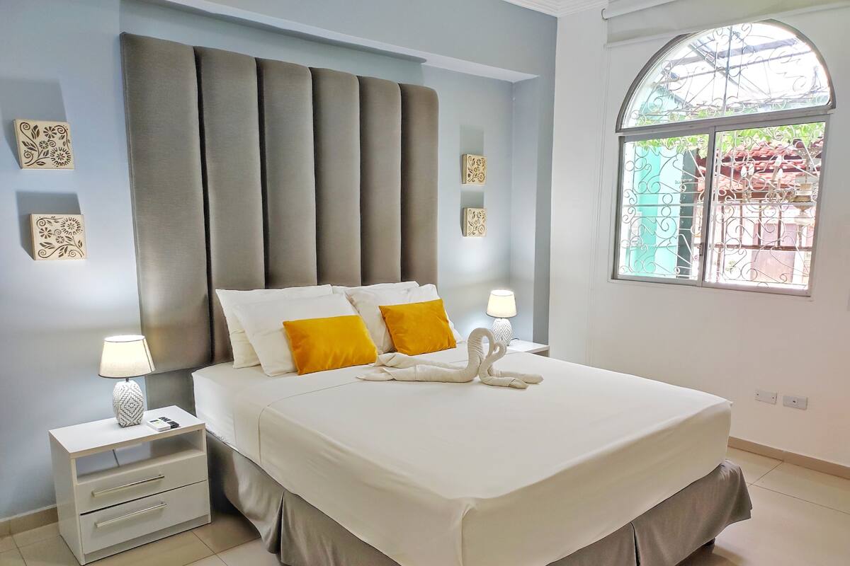 A neatly made bed with white and yellow pillows, a decorative swan pillow, and a grey upholstered headboard. There are two bedside tables with lamps, and a window with a metal grill.