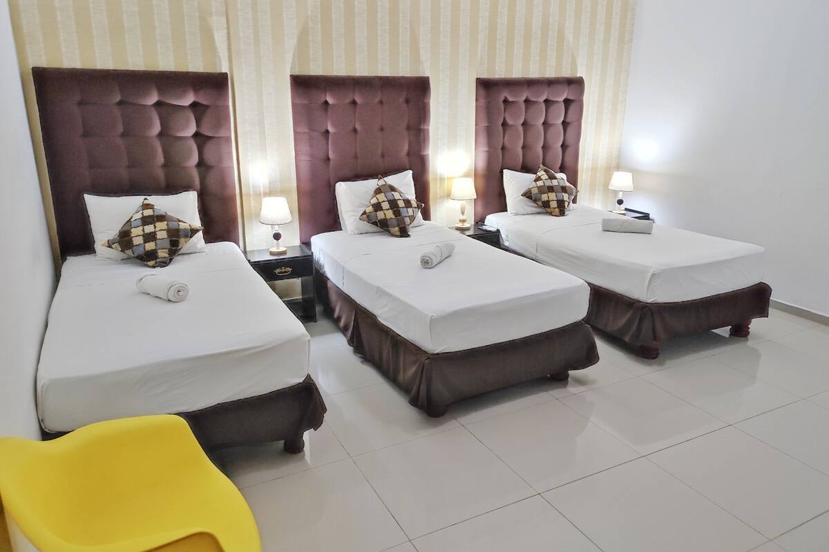 A hotel room with three single beds, each with a headboard, bedside tables, and lamps.