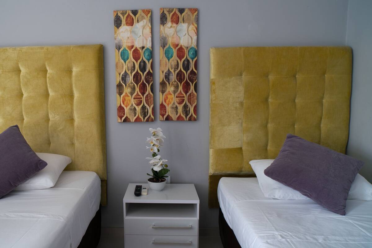 A hotel room with two beds, each with a tufted headboard, white bedding, and purple pillows. There is a small bedside table with a remote control and a potted plant.