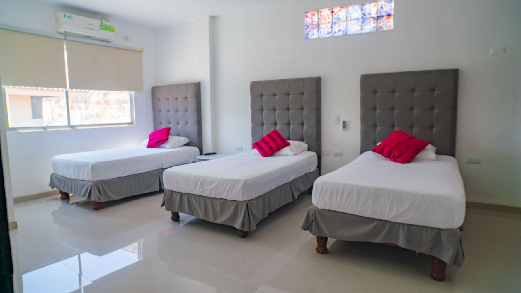 A hotel room with three beds, each with a grey headboard and white bedding, and a pink pillow.