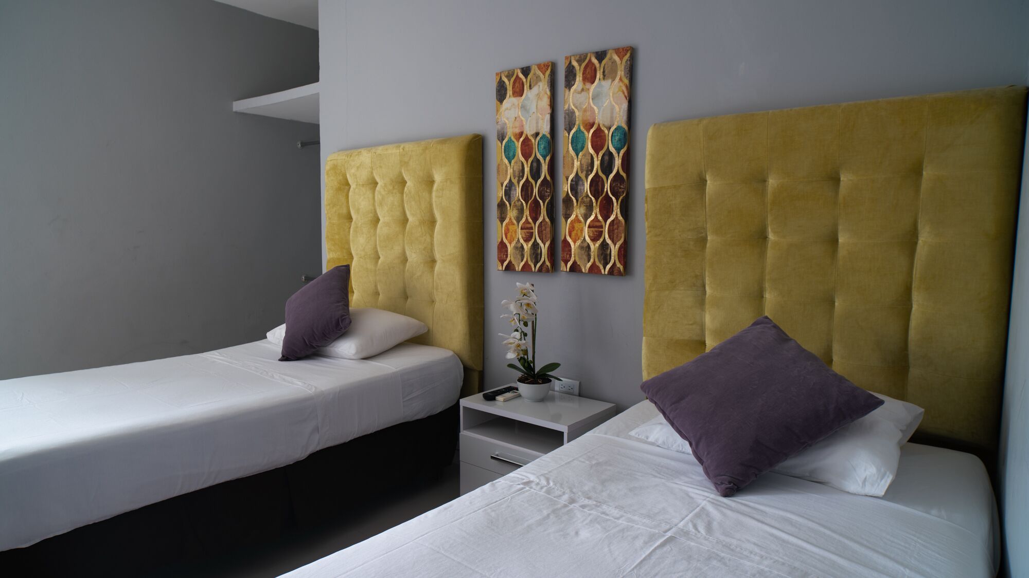 A hotel room with two single beds, a yellow tufted headboard, white bedding, a small bedside table with a remote control, and a potted orchid.