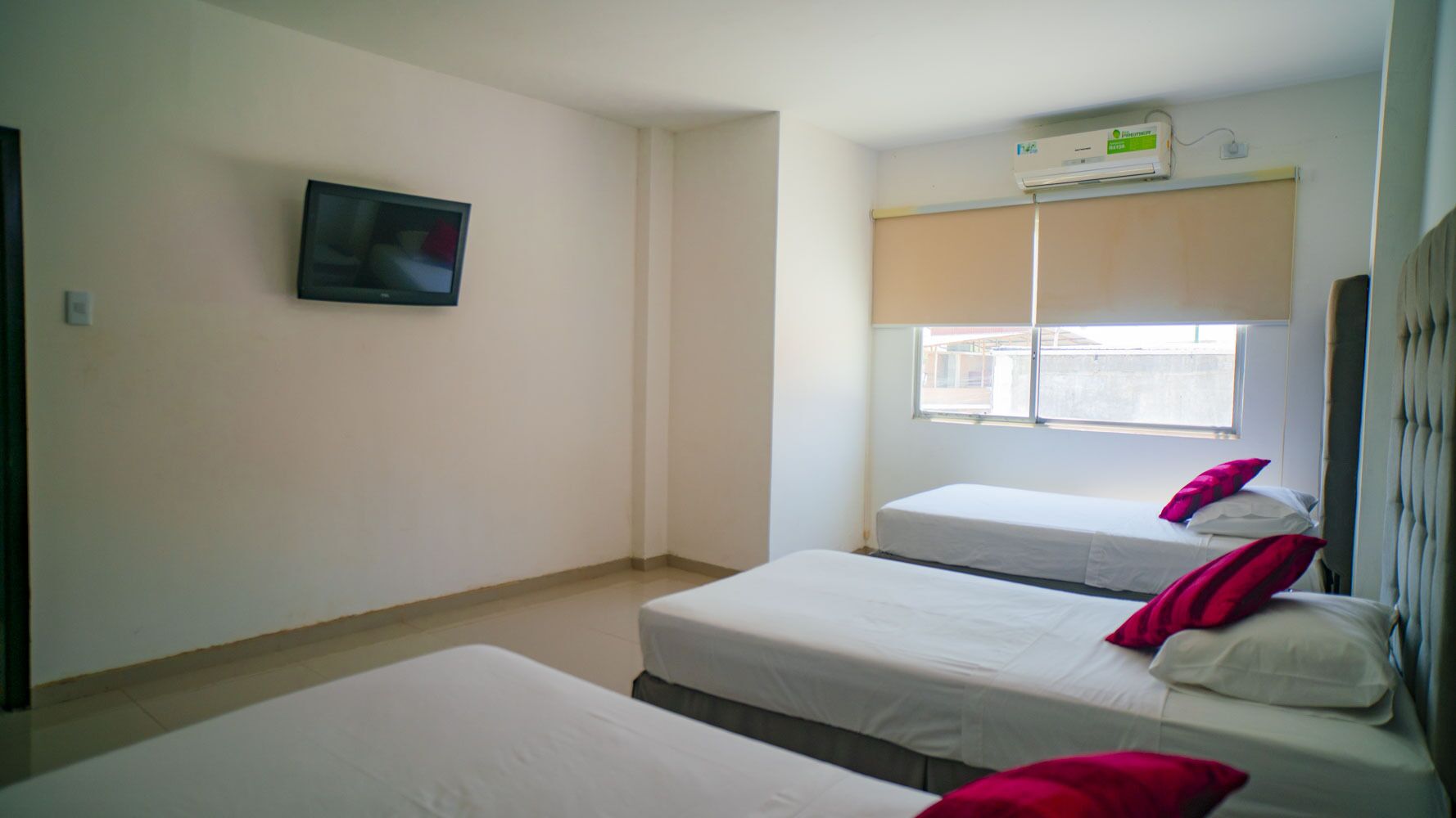 Una habitación de hotel con dos camas individuales, un televisor montado en la pared y una ventana con persiana color beige.