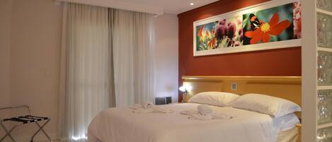 Una habitación de hotel con una cama grande, un cuadro de flores en la pared, una ventana con cortinas y un radiador.
