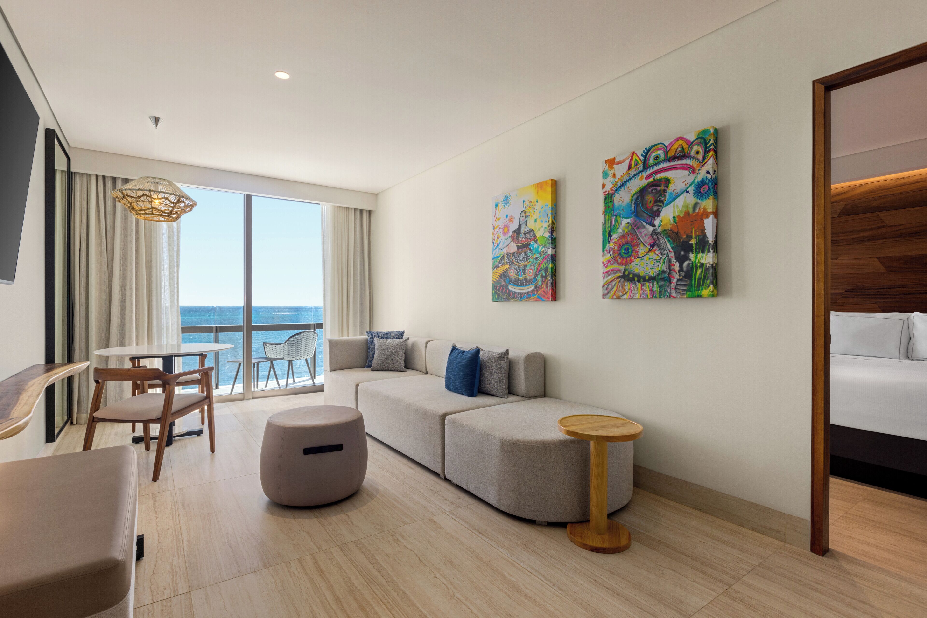 A modern hotel room with a large window, a sofa, a round table, and two artworks on the wall.