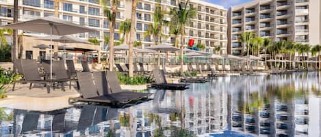 A hotel pool area with lounge chairs, umbrellas, and palm trees.