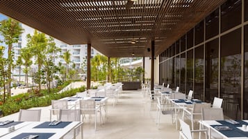 An outdoor patio area with white tables and chairs, a wooden ceiling, and large windows.
