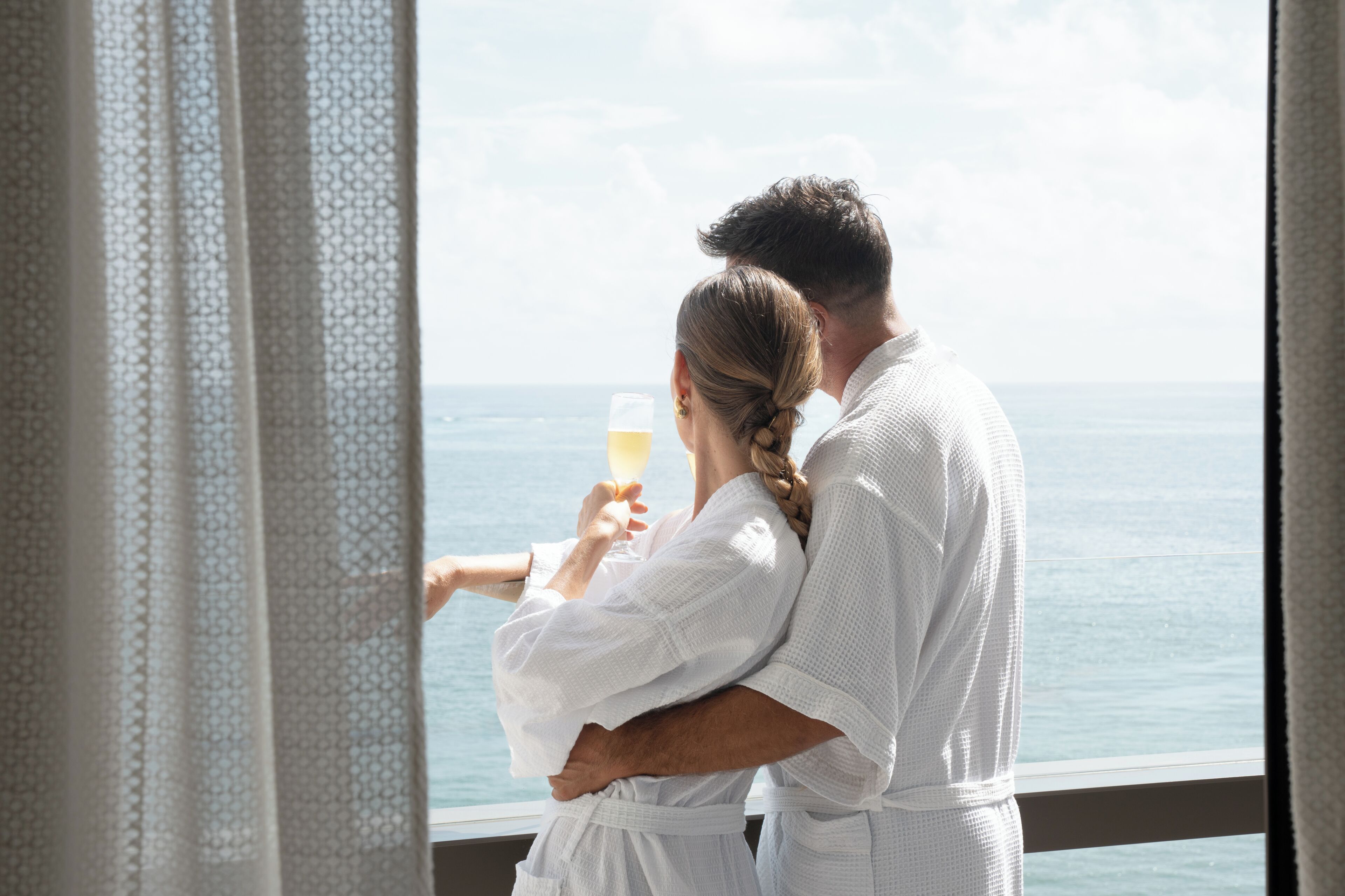 Una pareja vestida de blanco se abraza junto a una ventana con vistas al mar.
