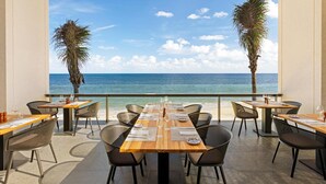 A restaurant with a long table set for dining, overlooking a beach and the sea.