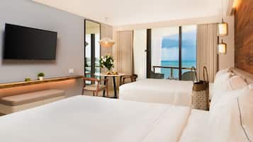 A hotel room with two beds, a TV, a balcony with a view of the sea, and a dining area with a table and chairs.