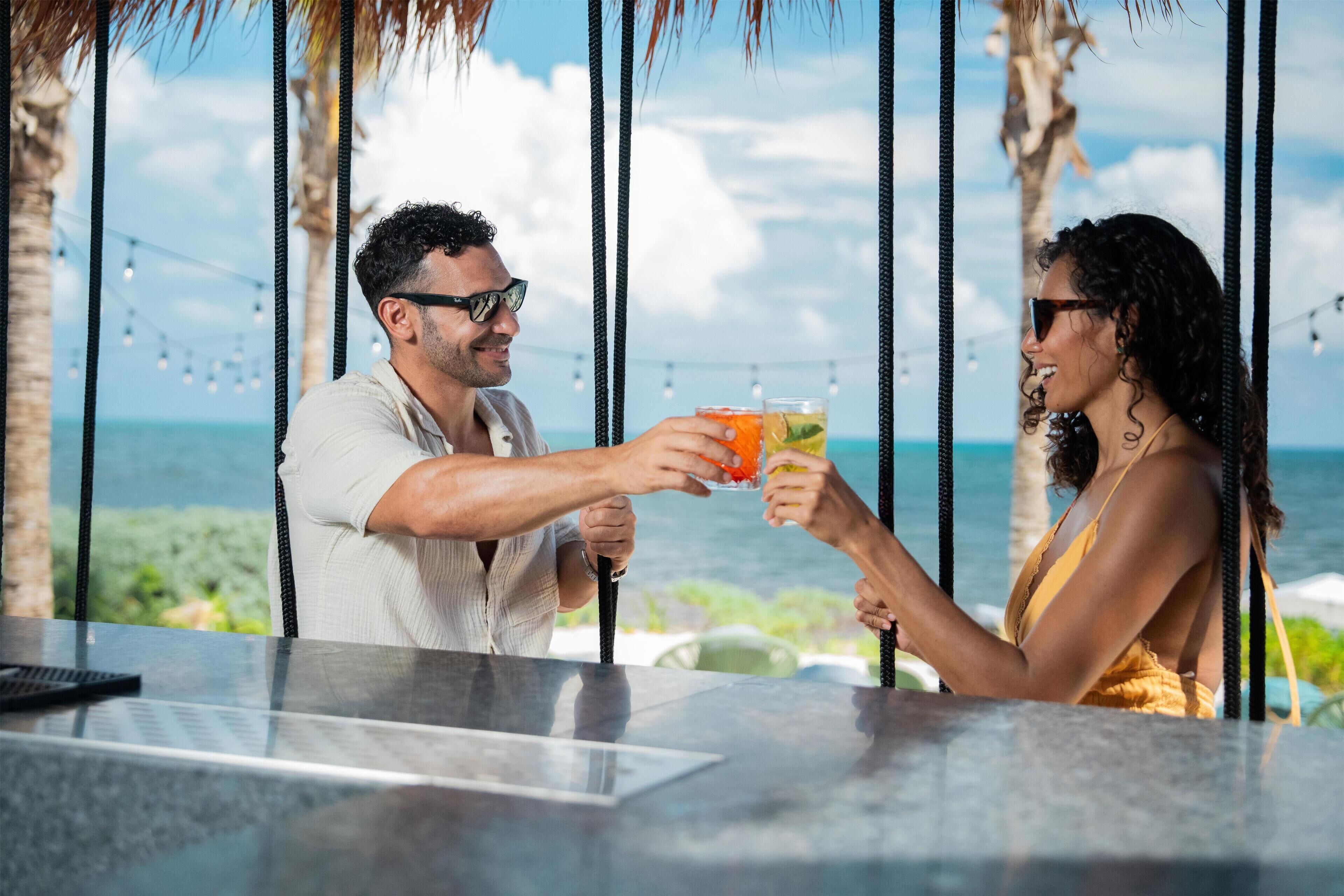 Una pareja brindando con bebidas en un bar con vista al océano y palmeras.