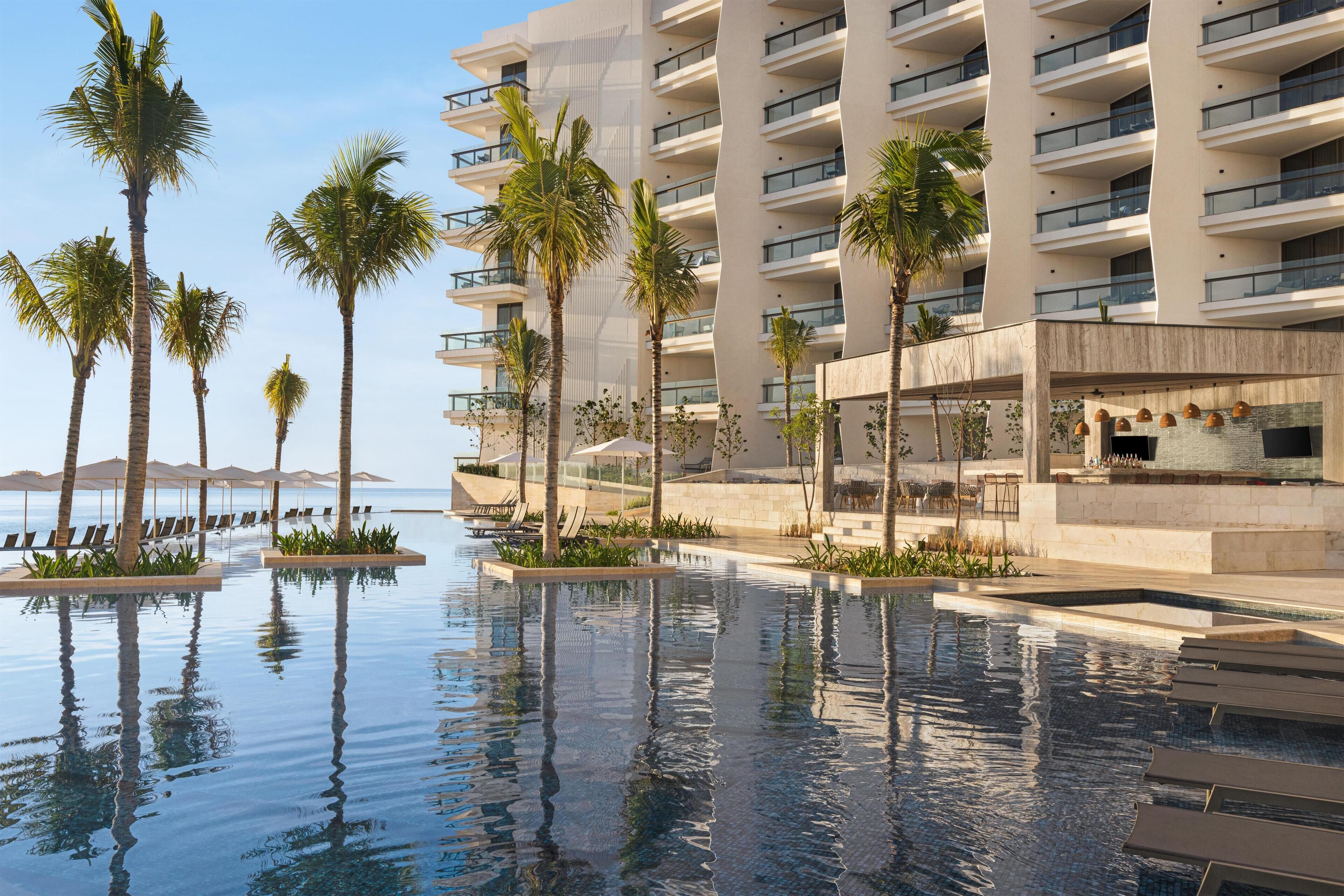 A modern hotel with a pool, palm trees, and a view of the ocean.