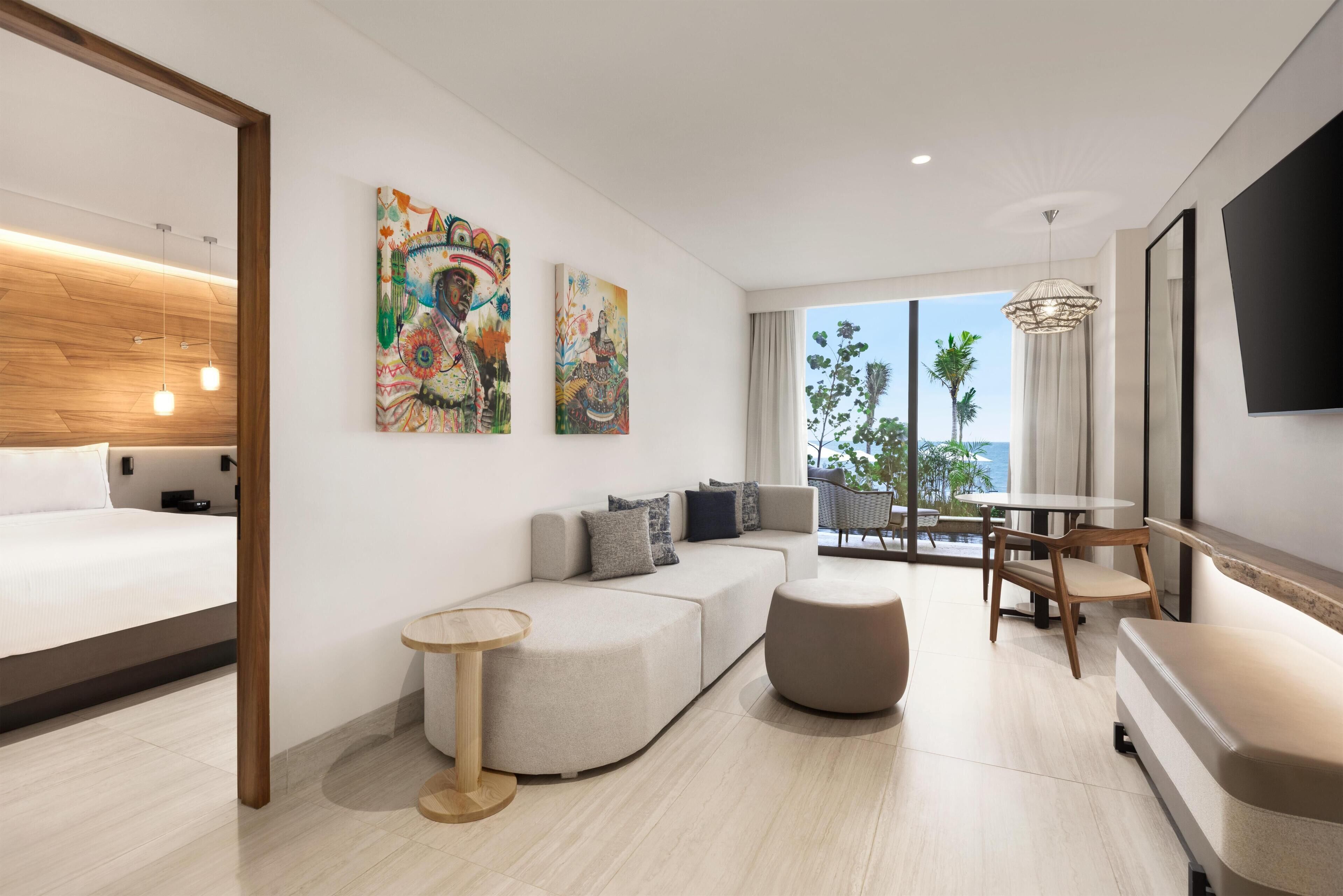 A modern hotel room with a bed, sofa, ottoman, and a dining area with a table and chairs. There are two large paintings on the wall and a TV mounted on the wall.